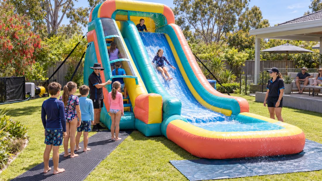 Inflatable water slide set up on grass in summer with children queuing at the steps.